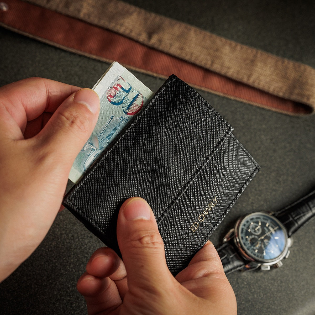 A close-up of a black textured bifold wallet being held by two hands, with a banknote partially inserted. A silver chronograph watch with a blue dial and a brown leather strap rest on a dark gray surface in the background.