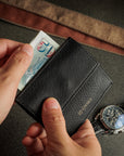 A close-up of a black textured bifold wallet being held by two hands, with a banknote partially inserted. A silver chronograph watch with a blue dial and a brown leather strap rest on a dark gray surface in the background.